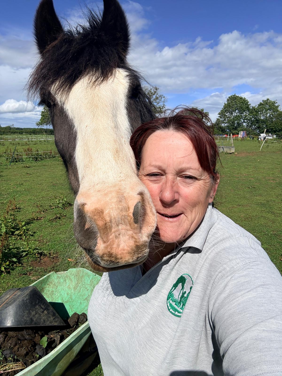 Brown and white horse with a white face, with his head against a ladies head who is staring at the screen, the lady has a green top on in a field