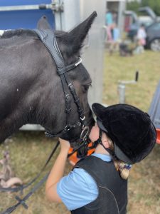 Black pony being kissed on the nose by a young girl in riding hat blonde hair