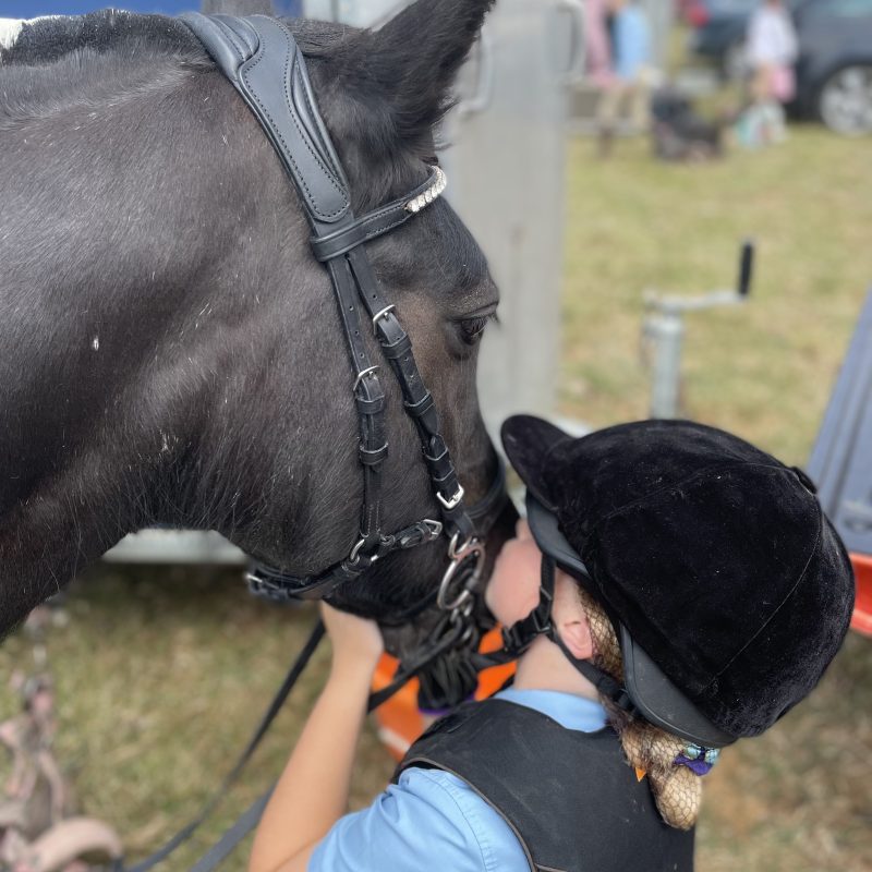 Black pony being kissed on the nose by a young girl in riding hat blonde hair