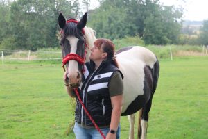 Lady wearing blue jeans, black and white gilet with red hair standing next to a black and white horse wearing a red headcollar