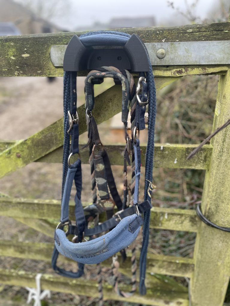 Wooden gate with metal hinge with black Bridle Buddy attached to the metal hinge with 2 blue headcollars hanging on it