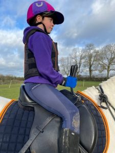 Young girl sitting on a horse with a purple and pink hat on with a purple top on with navy jodhpurs and blue gloves, holding Equihandee Bunny Ears whilst sitting on a horse in a black saddle