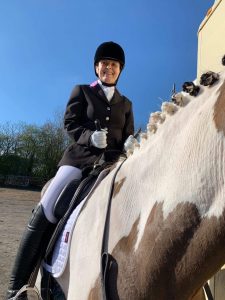 A lady smiling sitting on a horse looking down towards the ground holding Equihandee Bunny ears, wearing a black hat and riding jacket with white jodphurs and black boots sitting on a white and brown horse